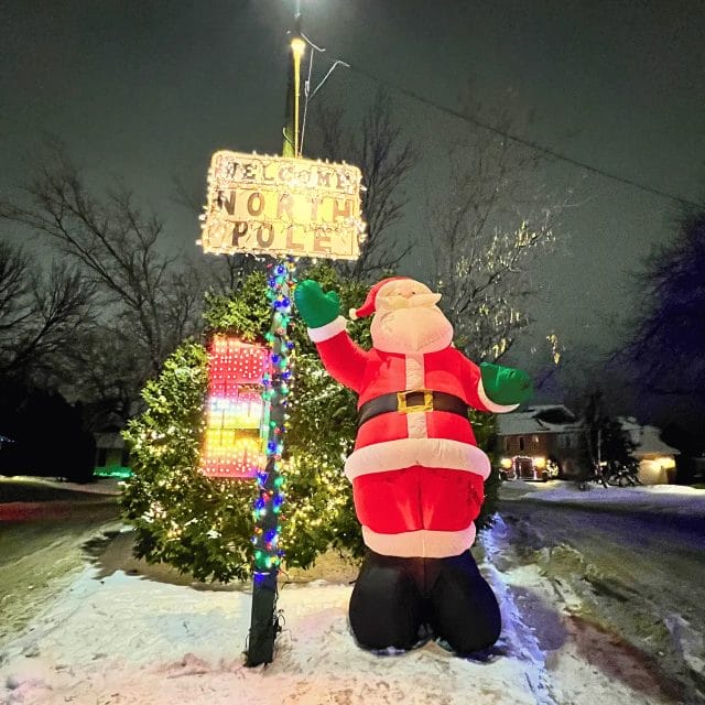 A Santa Claus standing in front of a pole with a sign that says, "Welcome to the North Pole" and "Ho ho ho!".