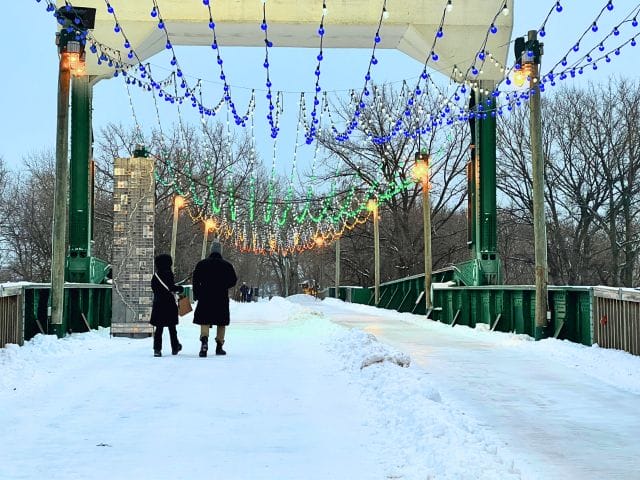 People walking under festive lights on the Historic Railway Bridge, which has a groomed skating path next to it.