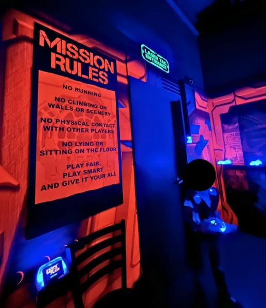 A young child (with his face blurred) standing in front of the entrance to the Laser Tag Game room in Uptown Alley. A board with the Mission Rules for the Laser Tag Game is next to the entrance.